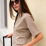 Woman in a beige outfit with sunglasses holding a suitcase against a neutral background