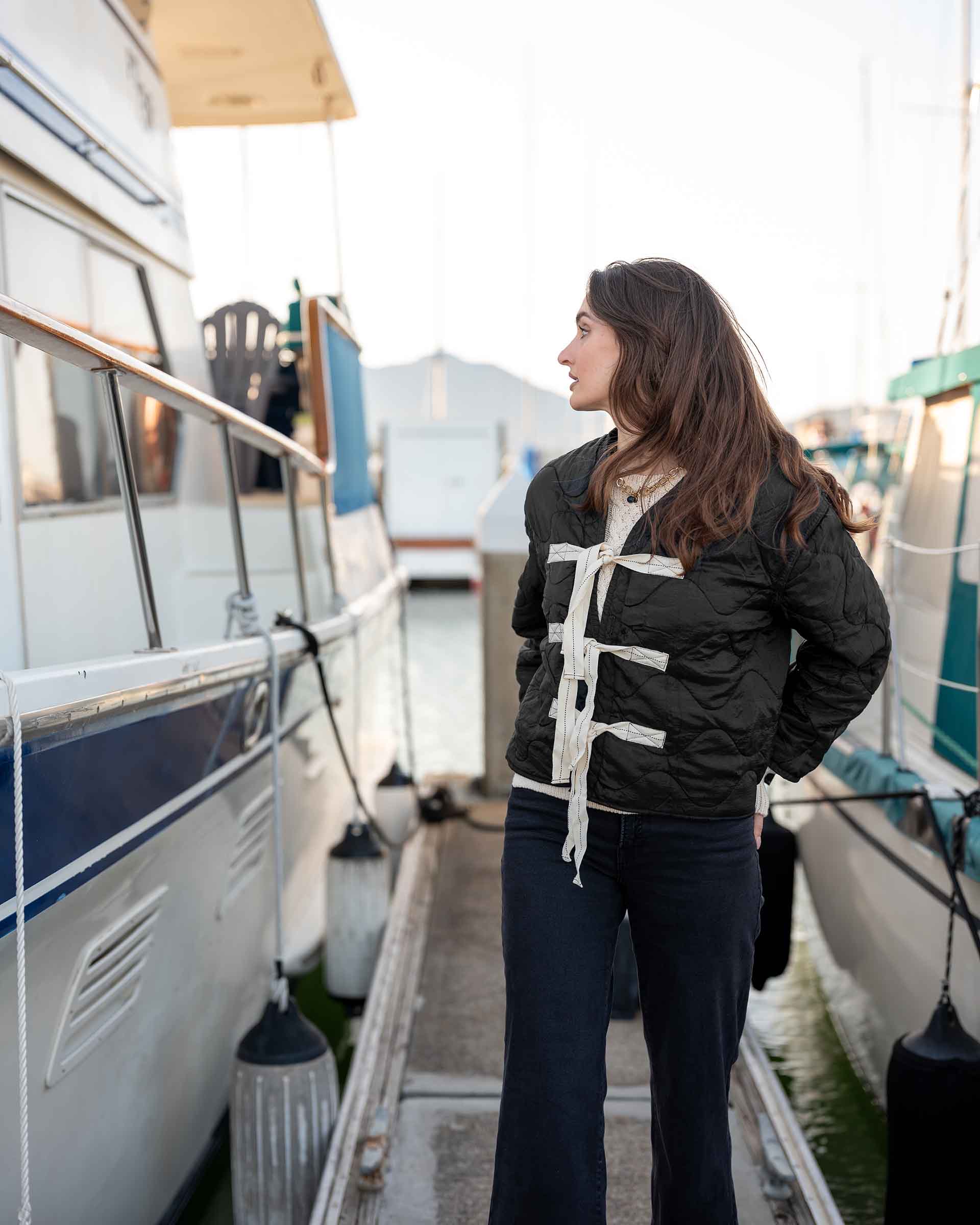 Woman standing on a dock next to boats, wearing a black jacket with lace-up front.