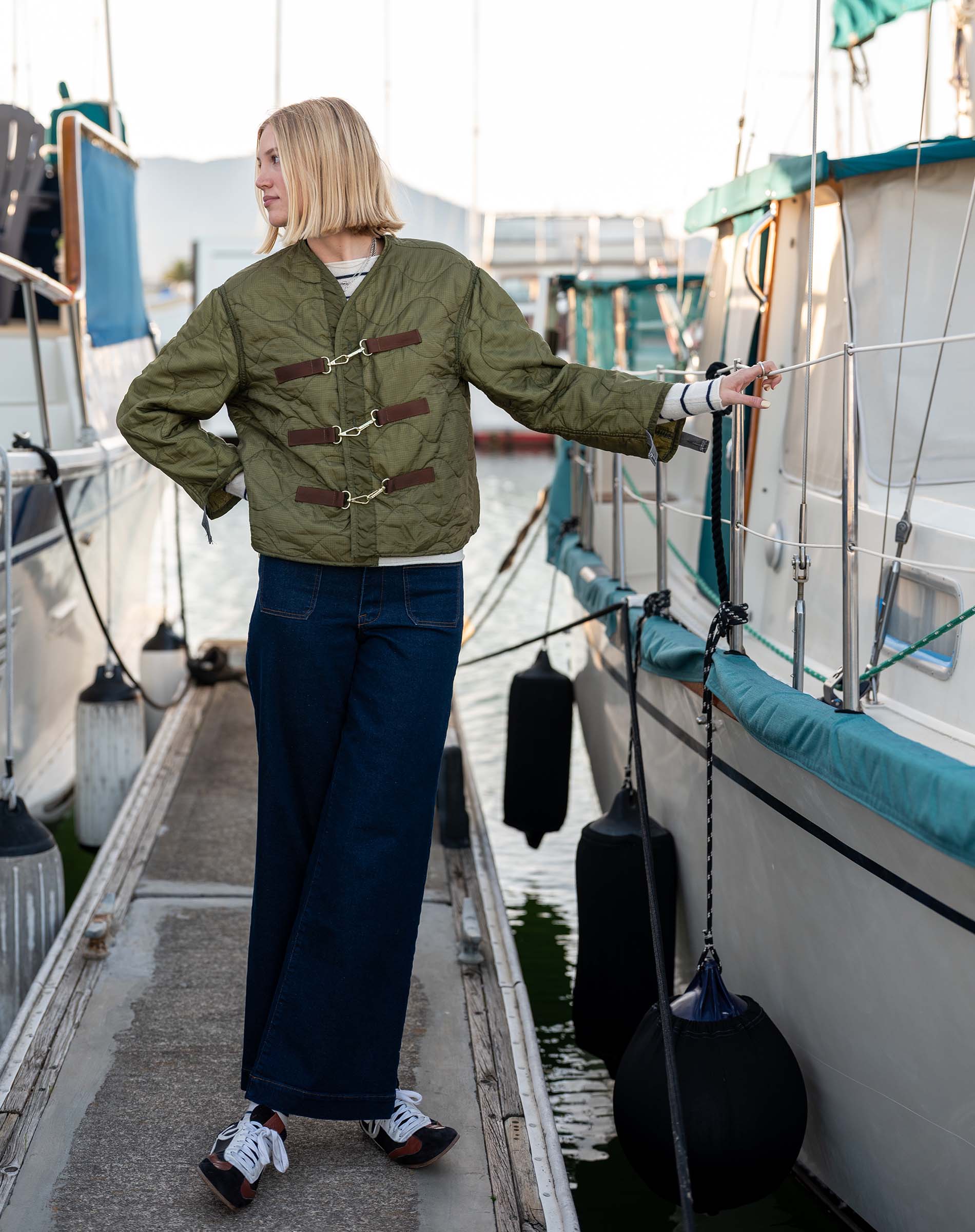 Person wearing a green jacket with brown buttons on a dock with boats in the background