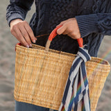 female holding straw basket with orange leather handles on the beach wearing a navy sweater