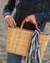 female holding straw basket with orange leather handles on the beach wearing a navy sweater