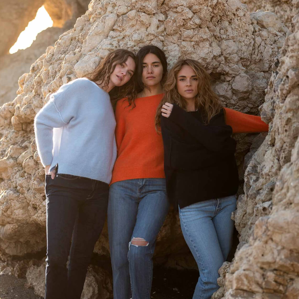 three women wearing blue, orange and navy sweater over jeans leaning against a rocky wall on the beach