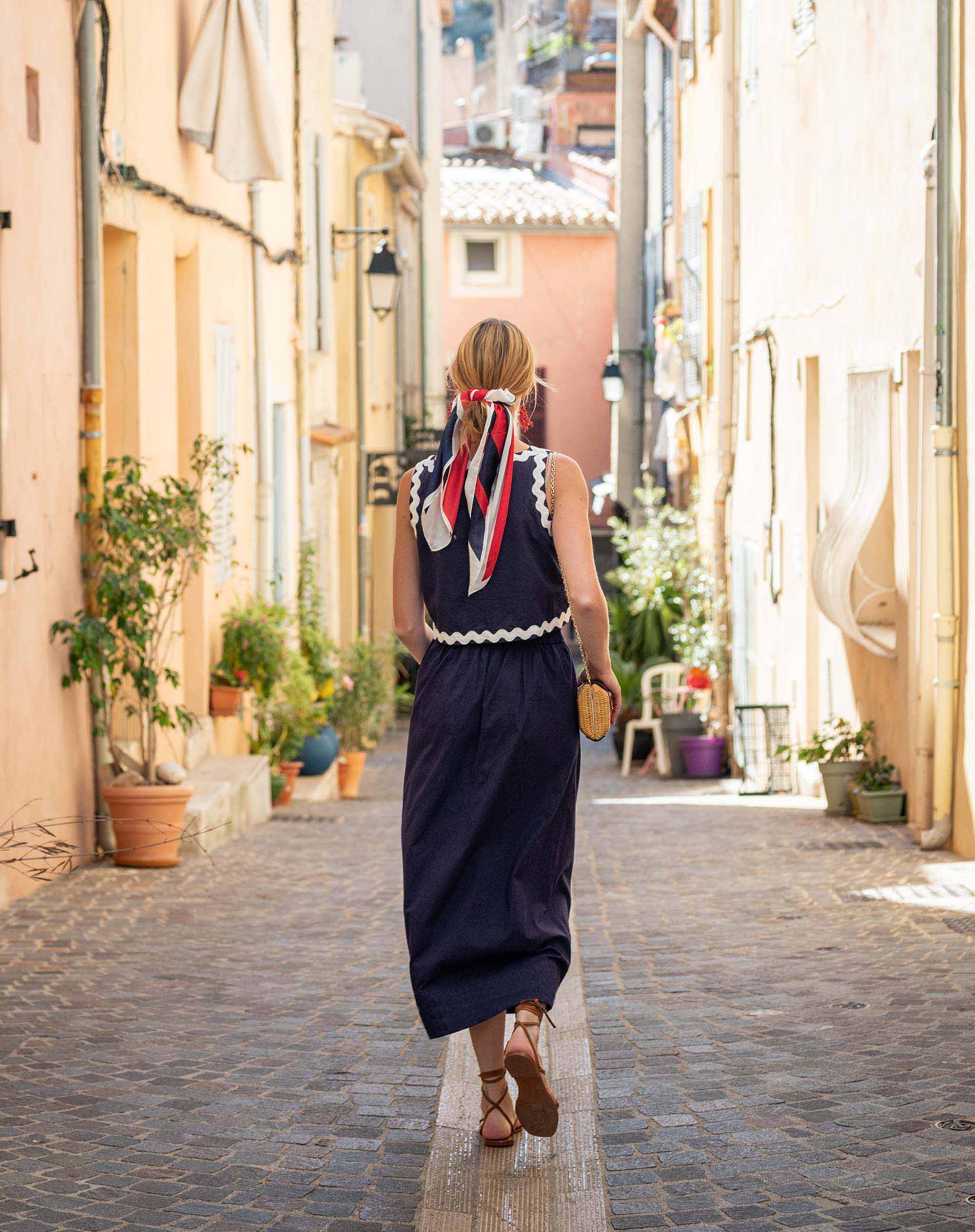 Woman in a navy dress with a red and white scarf walking down a narrow street in a European town.