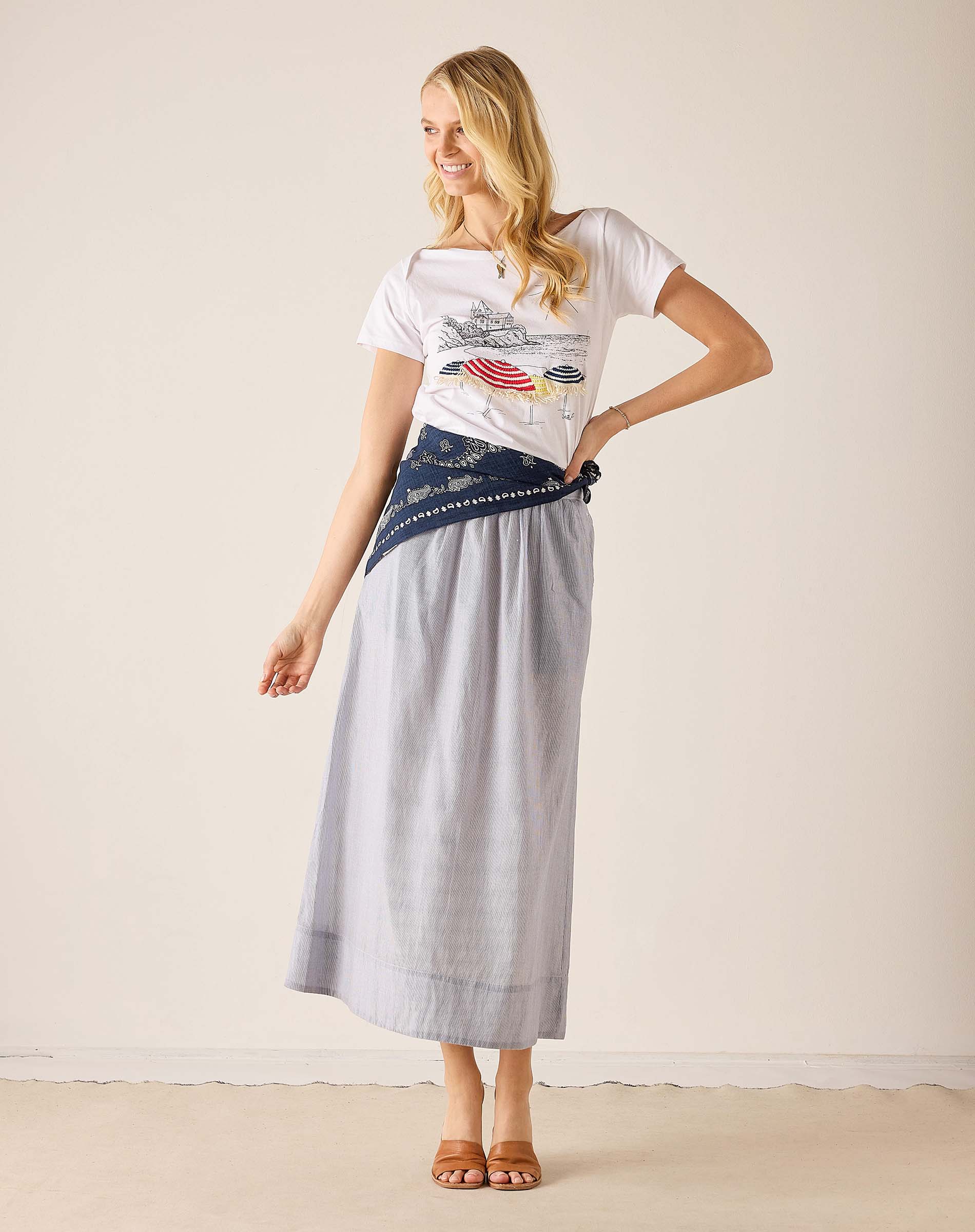 Woman wearing a white t-shirt with a graphic design and a long gray skirt on a plain background