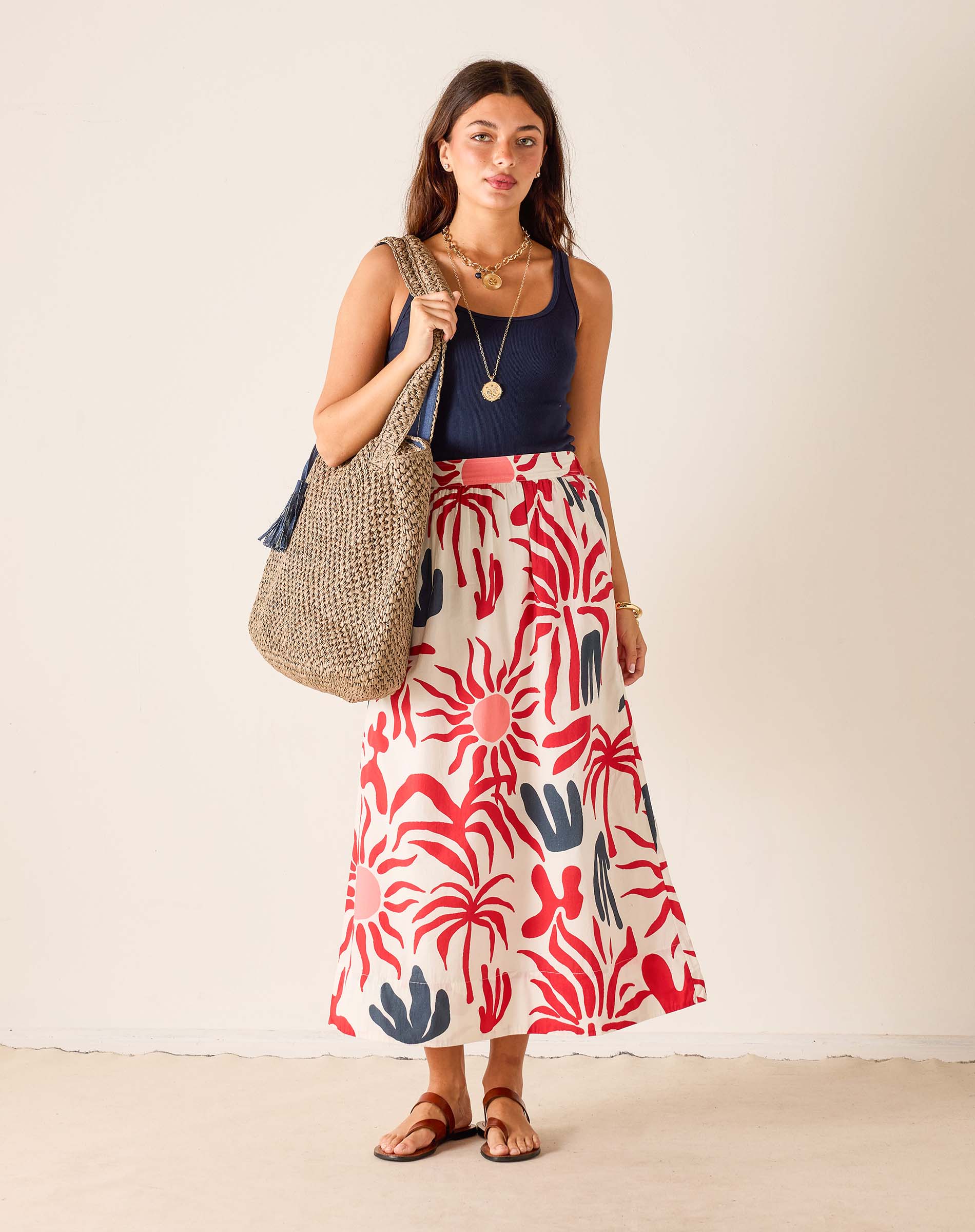 Woman wearing a red and blue patterned skirt holding a straw bag against a plain background