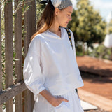 Woman in a white outfit with a striped headscarf standing outdoors near a wooden fence.
