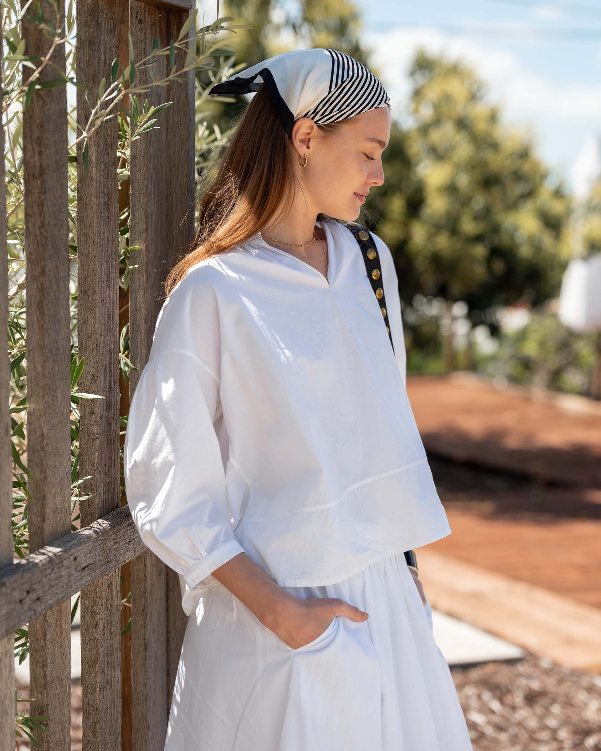 Woman in a white outfit with a striped headscarf standing outdoors near a wooden fence.