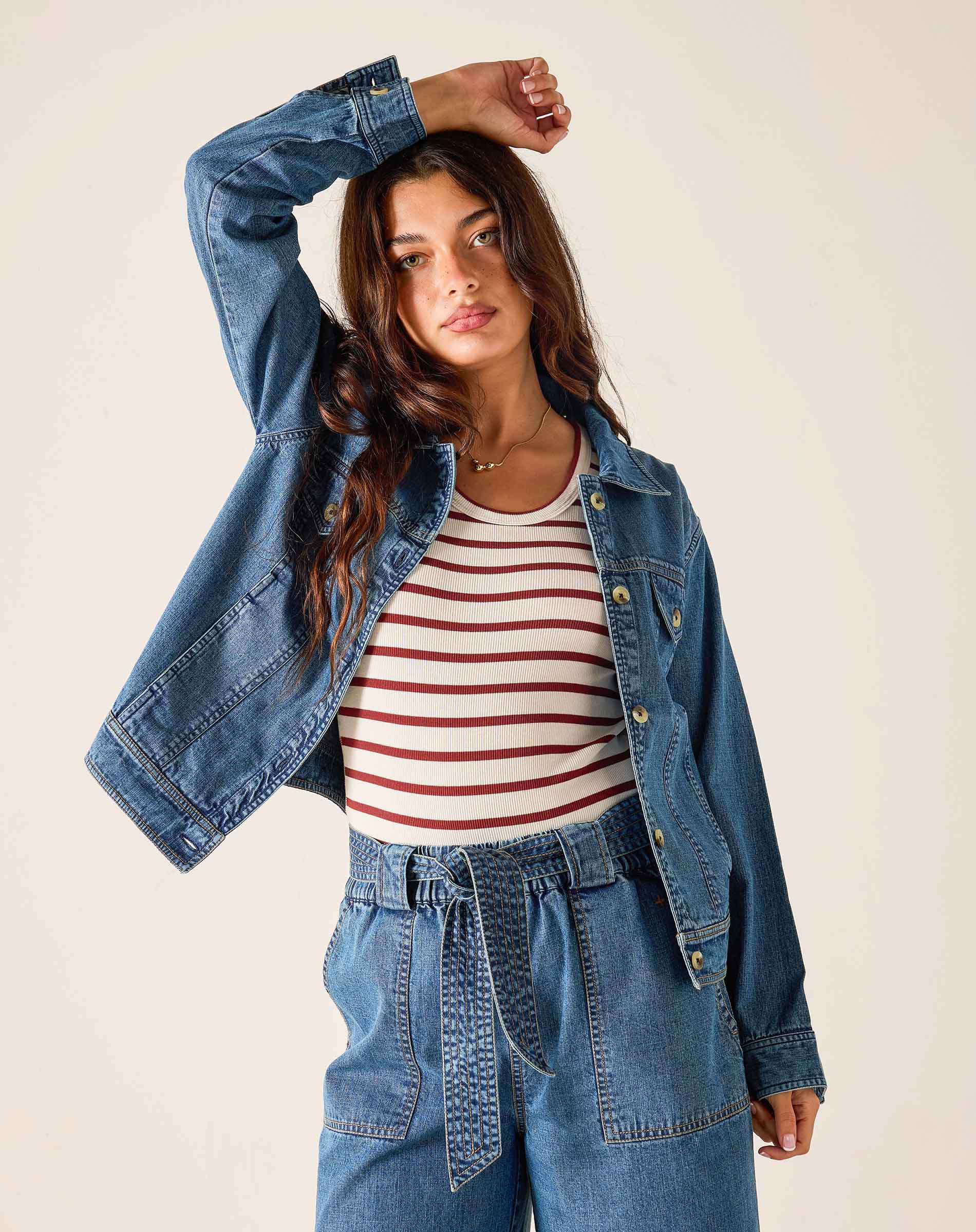 Woman wearing a denim jacket, striped shirt, and blue pants on a plain background