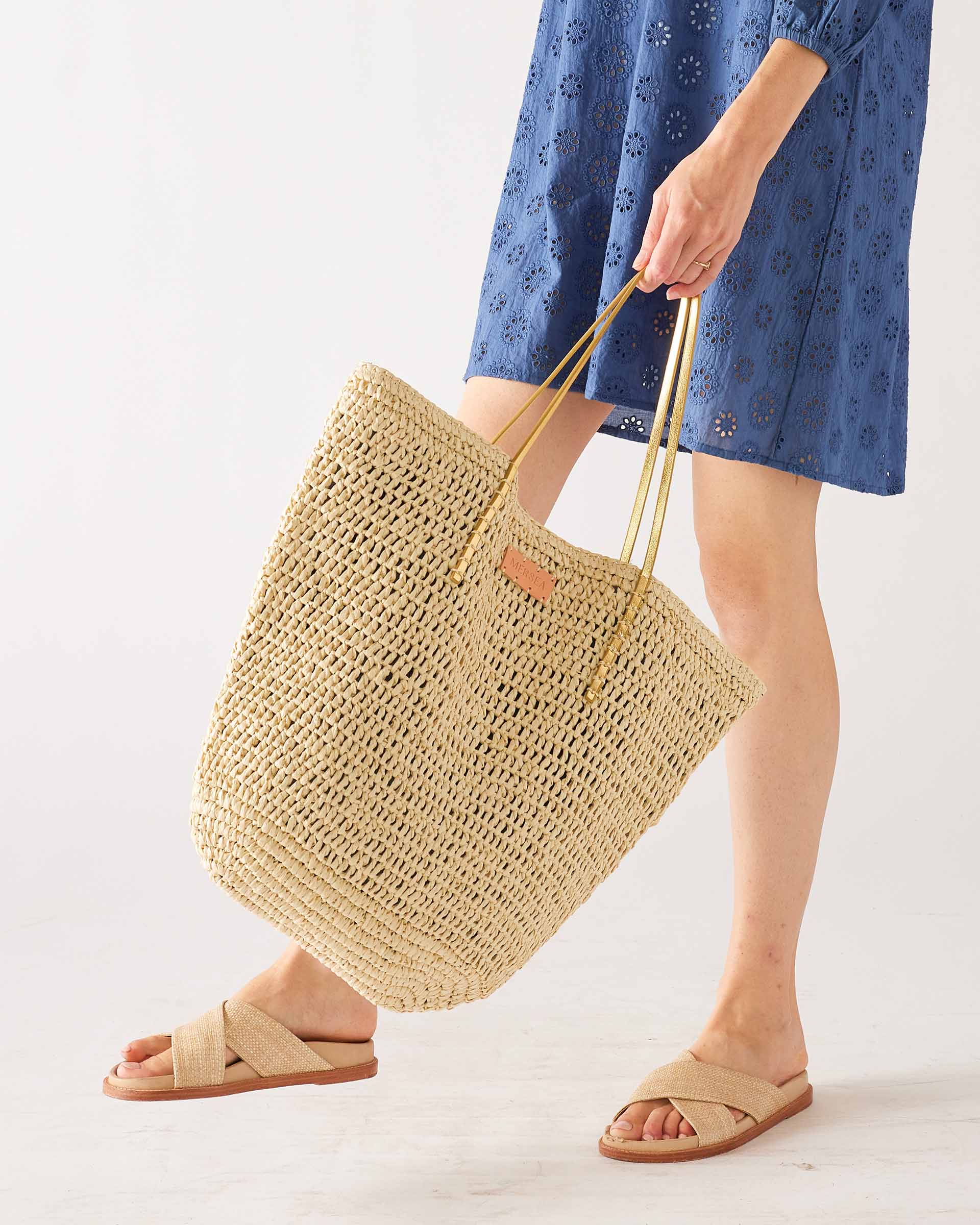 woman in blue dress holding sun chaser straw tote with sandals on