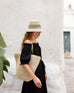 woman against a white wall in a black dress holding sun chaser straw tote