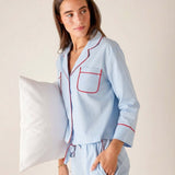 Woman wearing light blue pajamas with red piping, holding a pillow against a neutral background