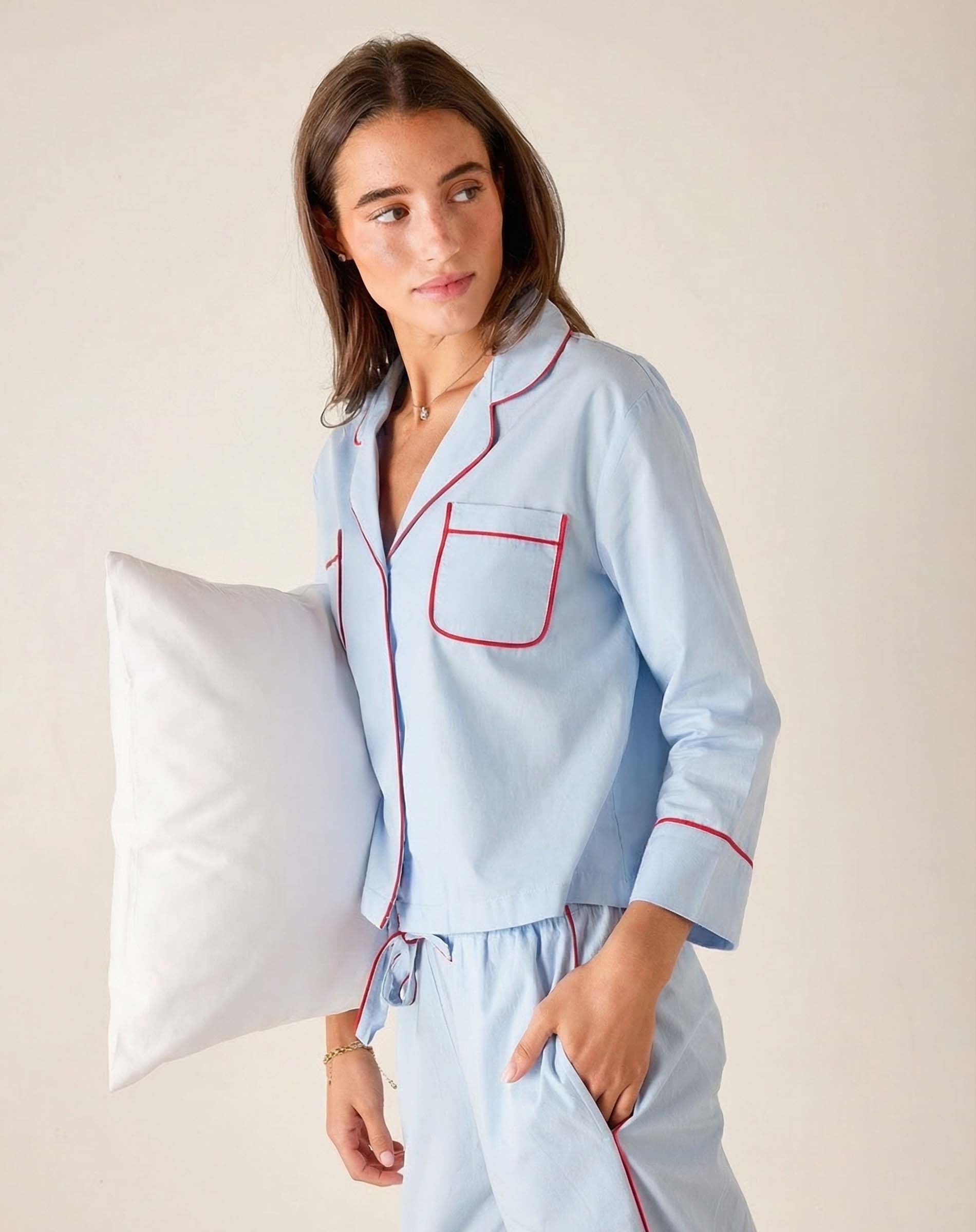 Woman wearing light blue pajamas with red piping, holding a pillow against a neutral background