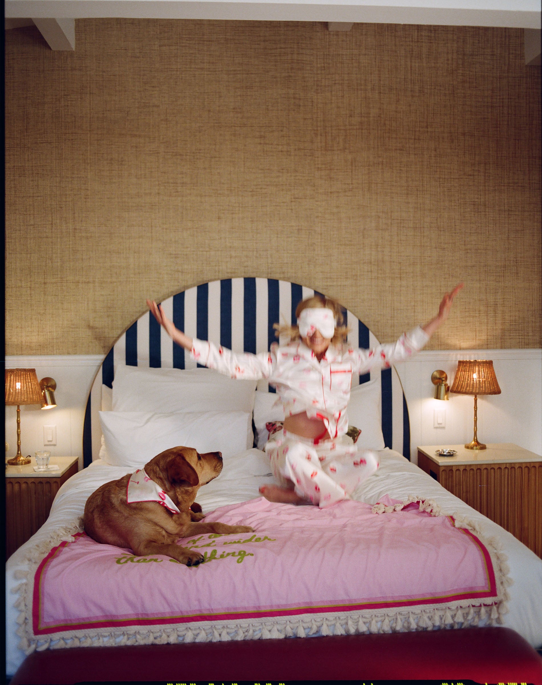 Woman in a pink robe sitting on a bed with a dog, in a bedroom setting.