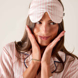 Woman wearing a pink checkered sleep mask and pajamas, posing with hands on face.