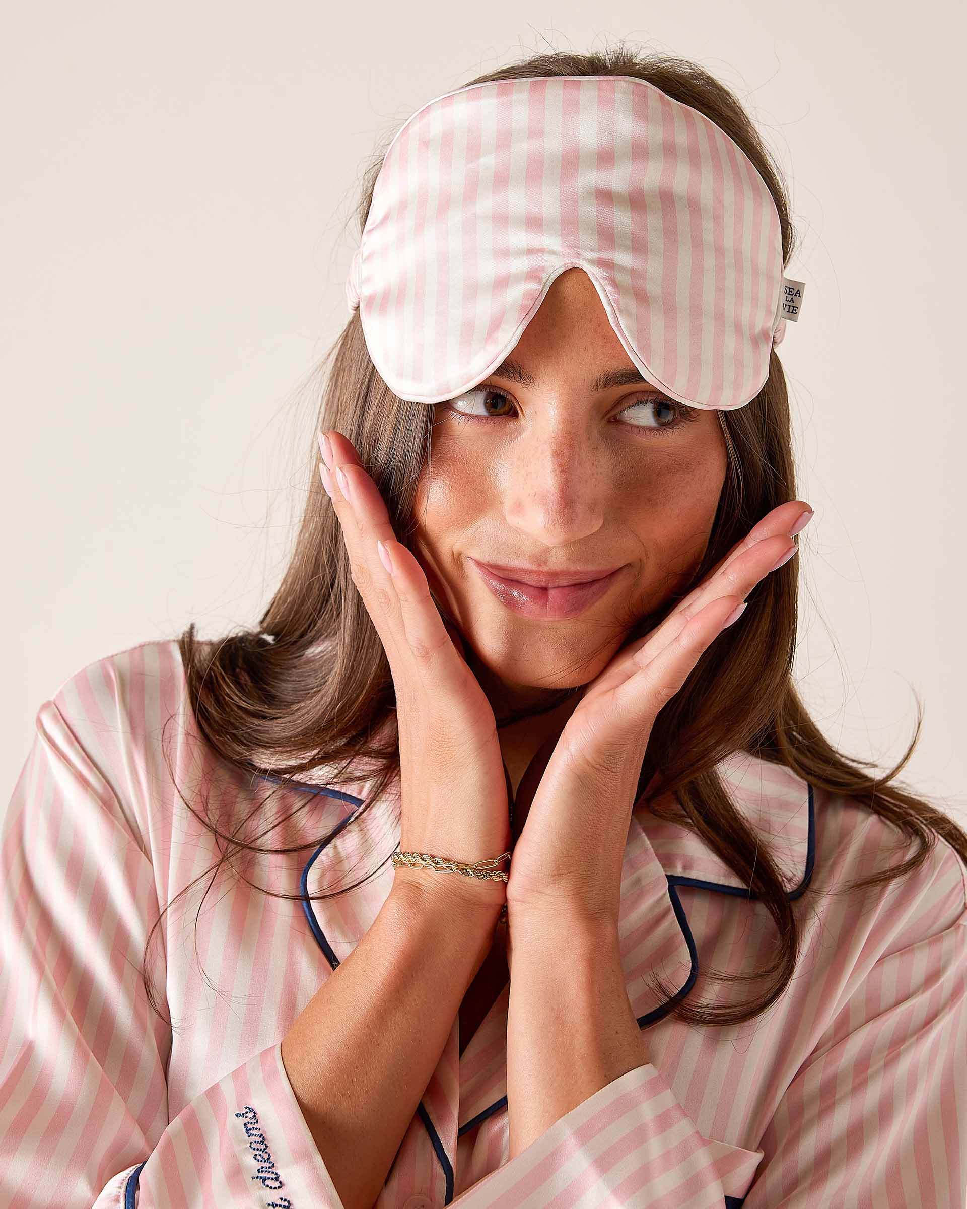 Woman wearing a pink checkered sleep mask and pajamas, posing with hands on face.