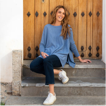 women wearing blue catalina sweater with split sides over travel jeans and white sneakers sitting on steps
