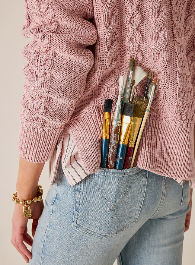 Close up image of a woman wearing a pink cable-knit sweater and paint brushes in back pocket
