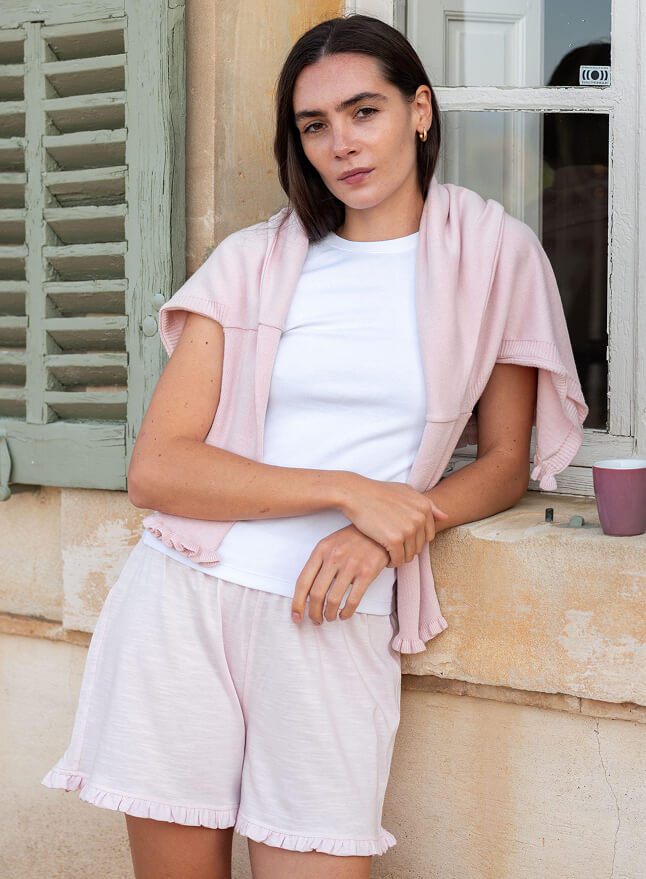 Woman wearing a light pink cardigan, white shirt, and shorts sitting on a window sill.