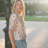 Woman wearing a short-sleeved, cream-colored shirt with a floral blossom and berry print and light-wash denim jeans.