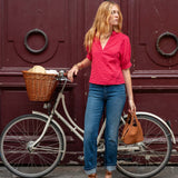 Woman in a red shirt and blue jeans standing next to a bicycle with a basket against a red door.