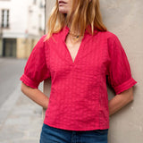Woman wearing a red blouse and blue jeans against a neutral wall.