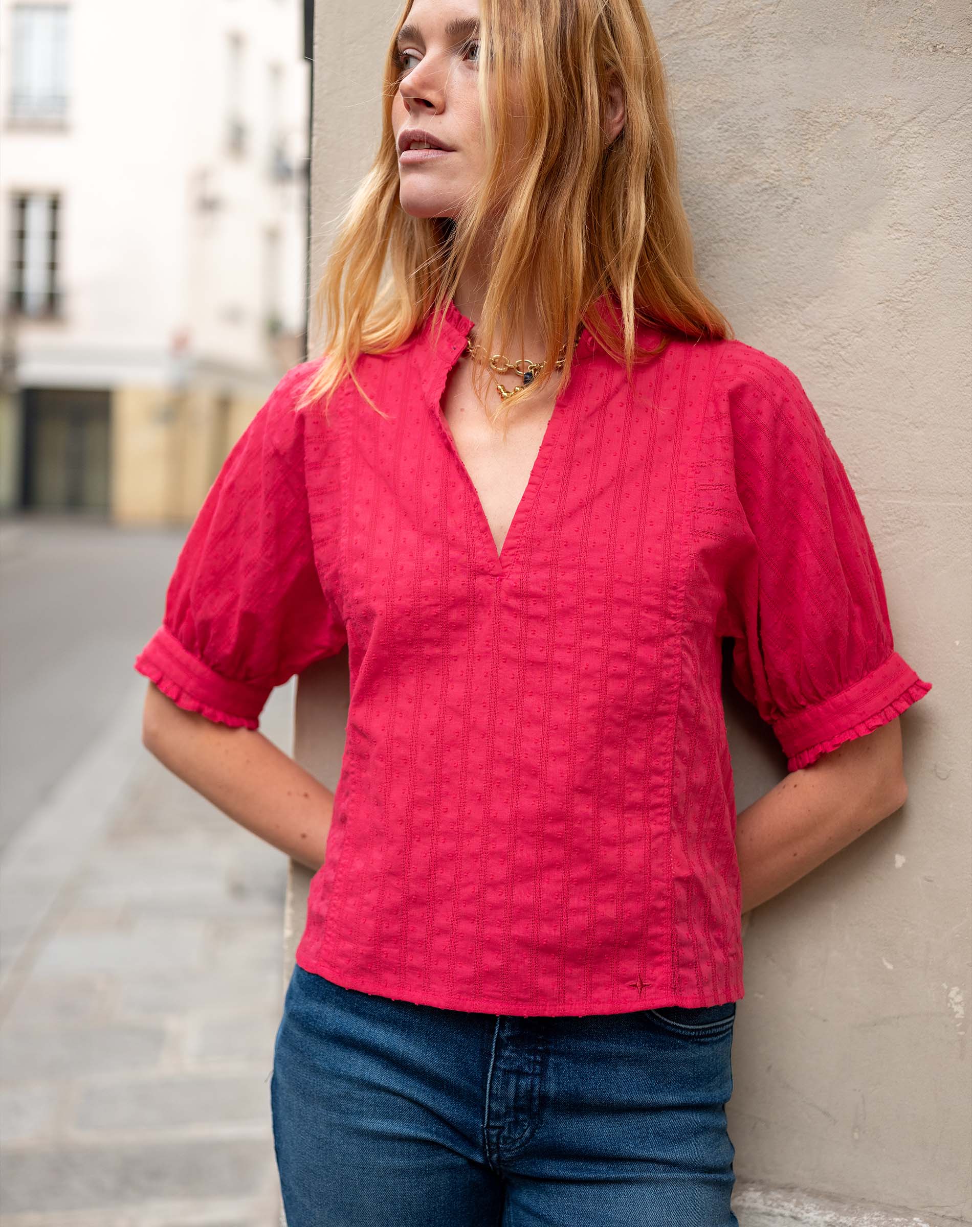Woman wearing a red blouse and blue jeans against a neutral wall.