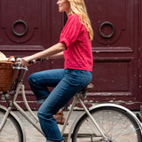 Woman in red shirt and blue jeans riding a bicycle with a basket against a red door.