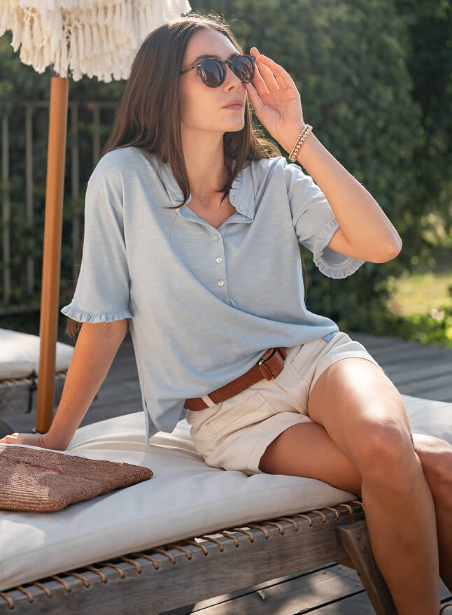 Woman sitting on a lounge chair outdoors, wearing sunglasses and a light blue shirt.