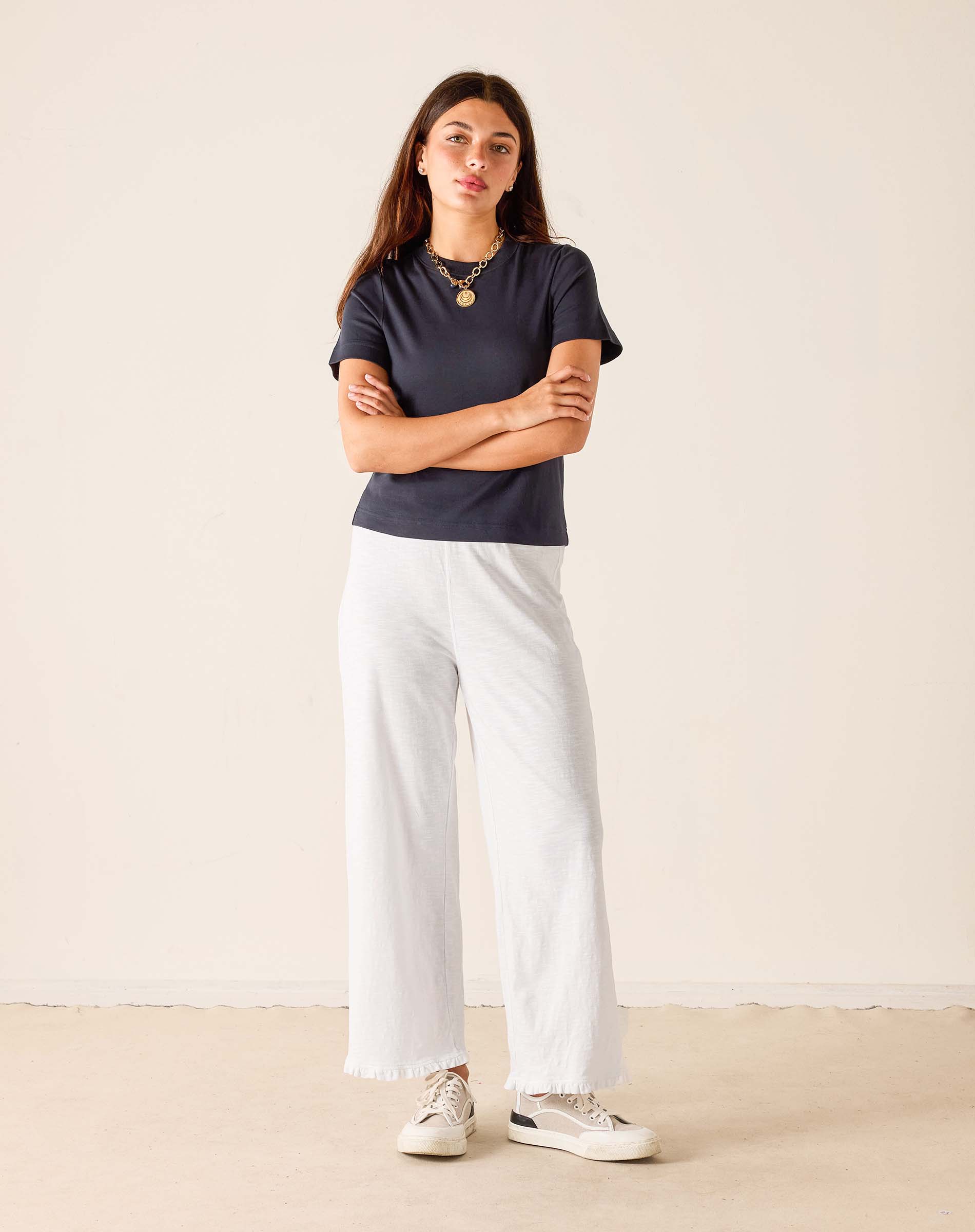 Woman wearing a navy t-shirt and white pants standing against a plain background