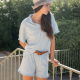 Woman wearing a light blue outfit with a hat, standing on a balcony with greenery in the background.