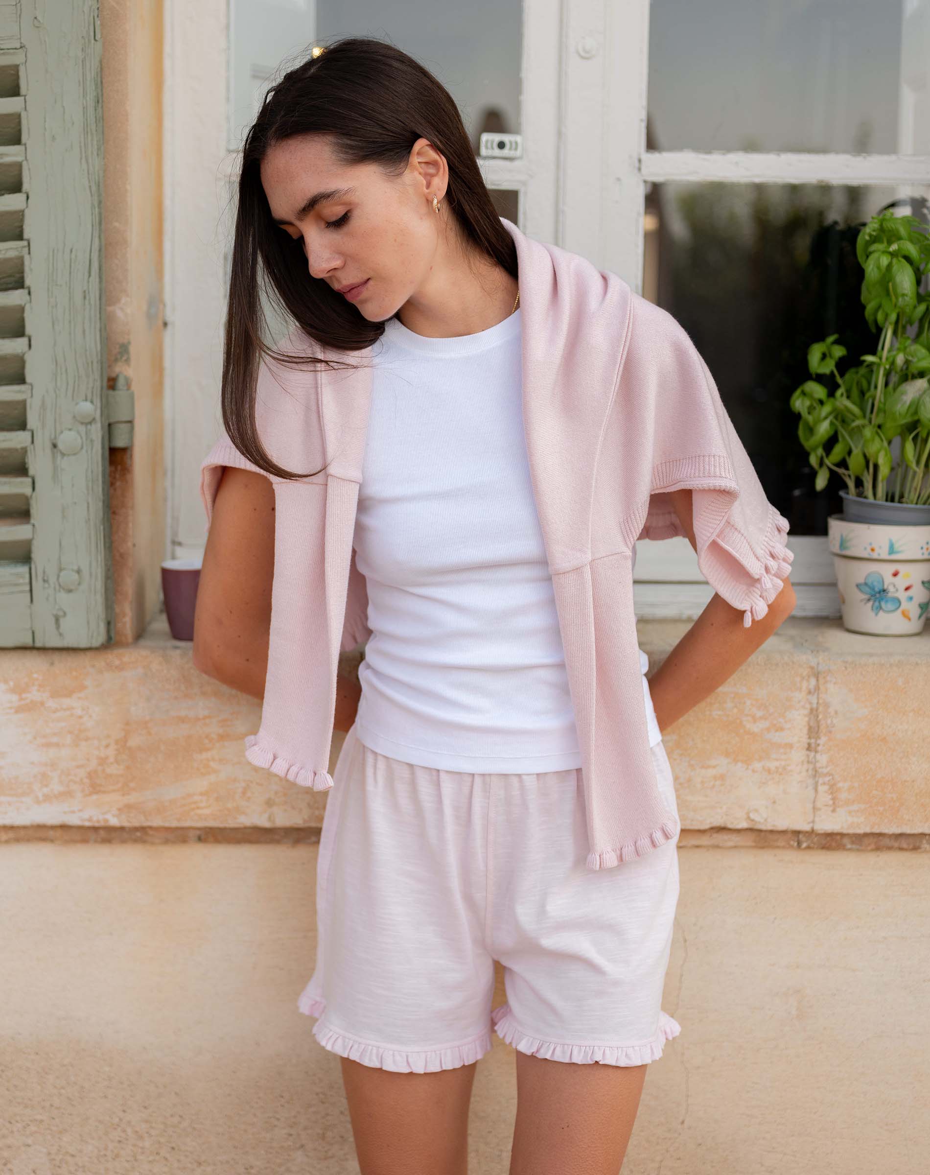 Woman wearing a light pink cardigan, white top, and shorts standing on a porch.