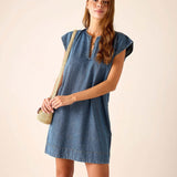 Woman wearing a blue denim dress holding a straw bag against a beige background