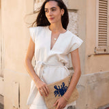 Woman in a white outfit holding a clutch with a blue crab design against a stone wall.