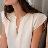 Woman wearing a white sleeveless top with a gold necklace, sitting against a neutral background.