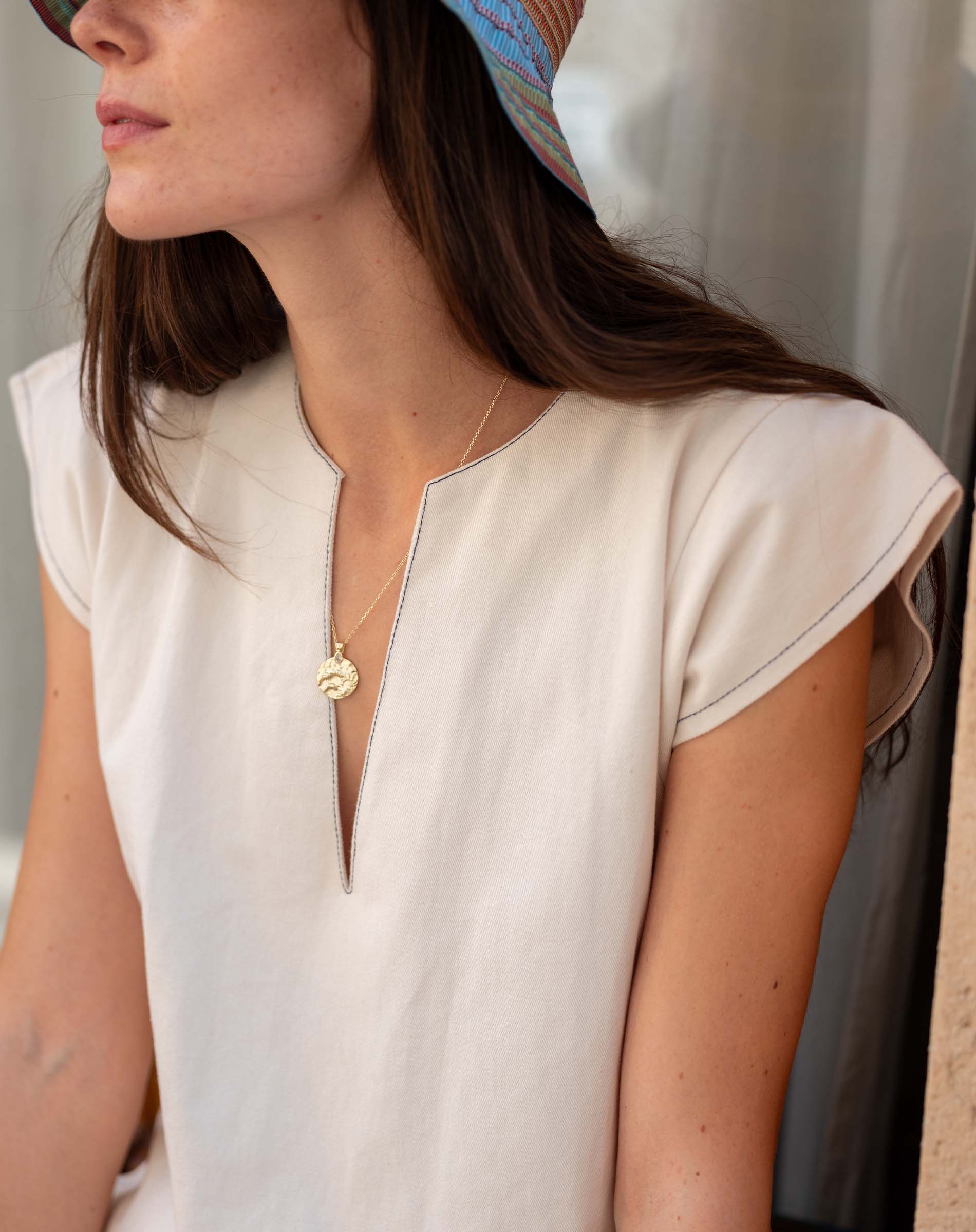 Woman wearing a white sleeveless top with a gold necklace, sitting against a neutral background.
