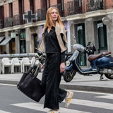 A woman in a black outfit and grey cardigan walking across a city street with a large tote bag.