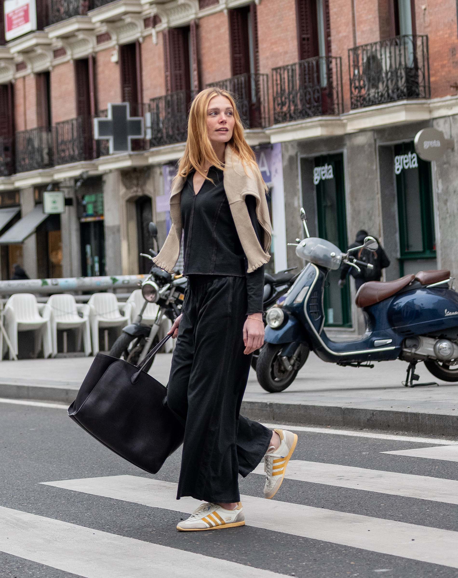 A woman in a black outfit and grey cardigan walking across a city street with a large tote bag.
