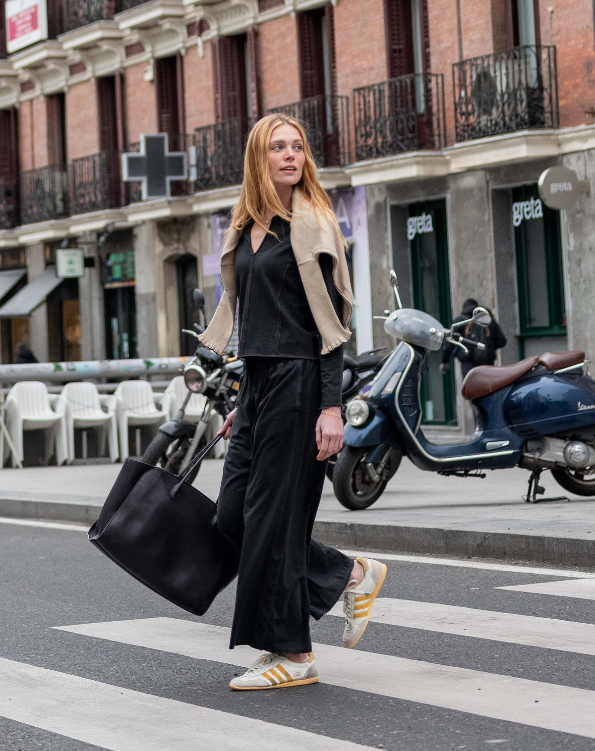 A woman in a black outfit and grey cardigan walking across a city street with a large tote bag.