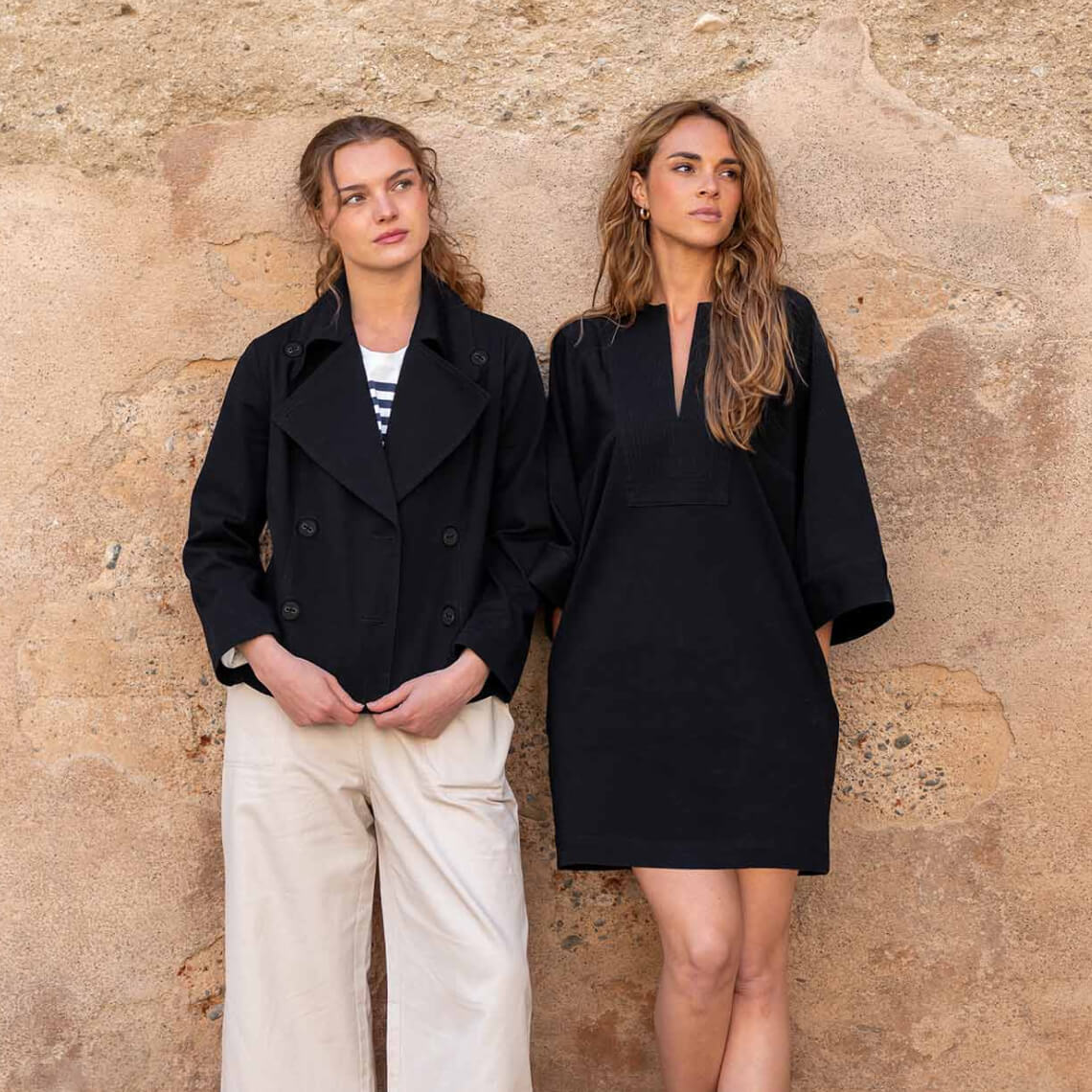 two females leaning against a brown wall wearing twill navy dress and a twill coat and pants