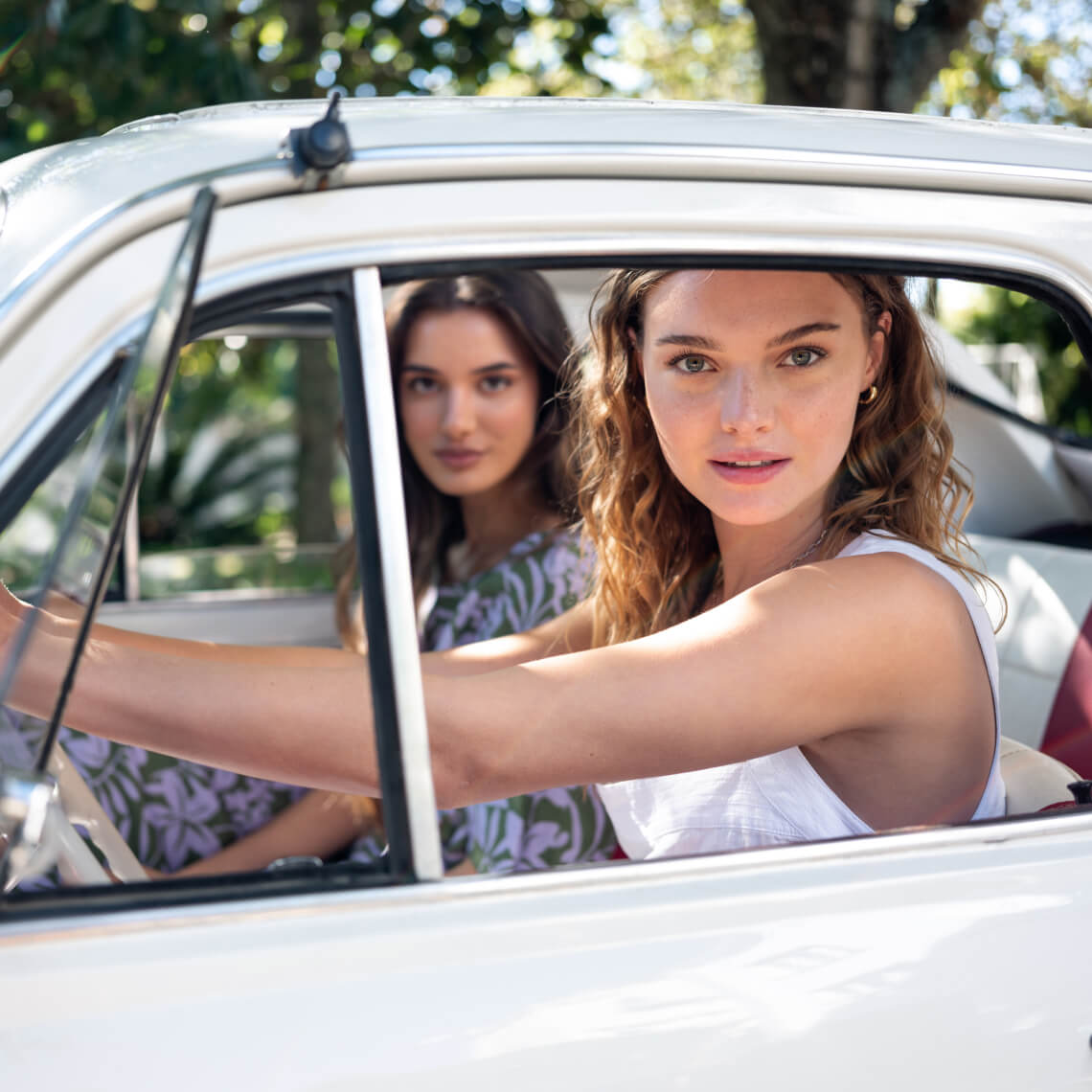 Two female sitting in a white car look at the camera