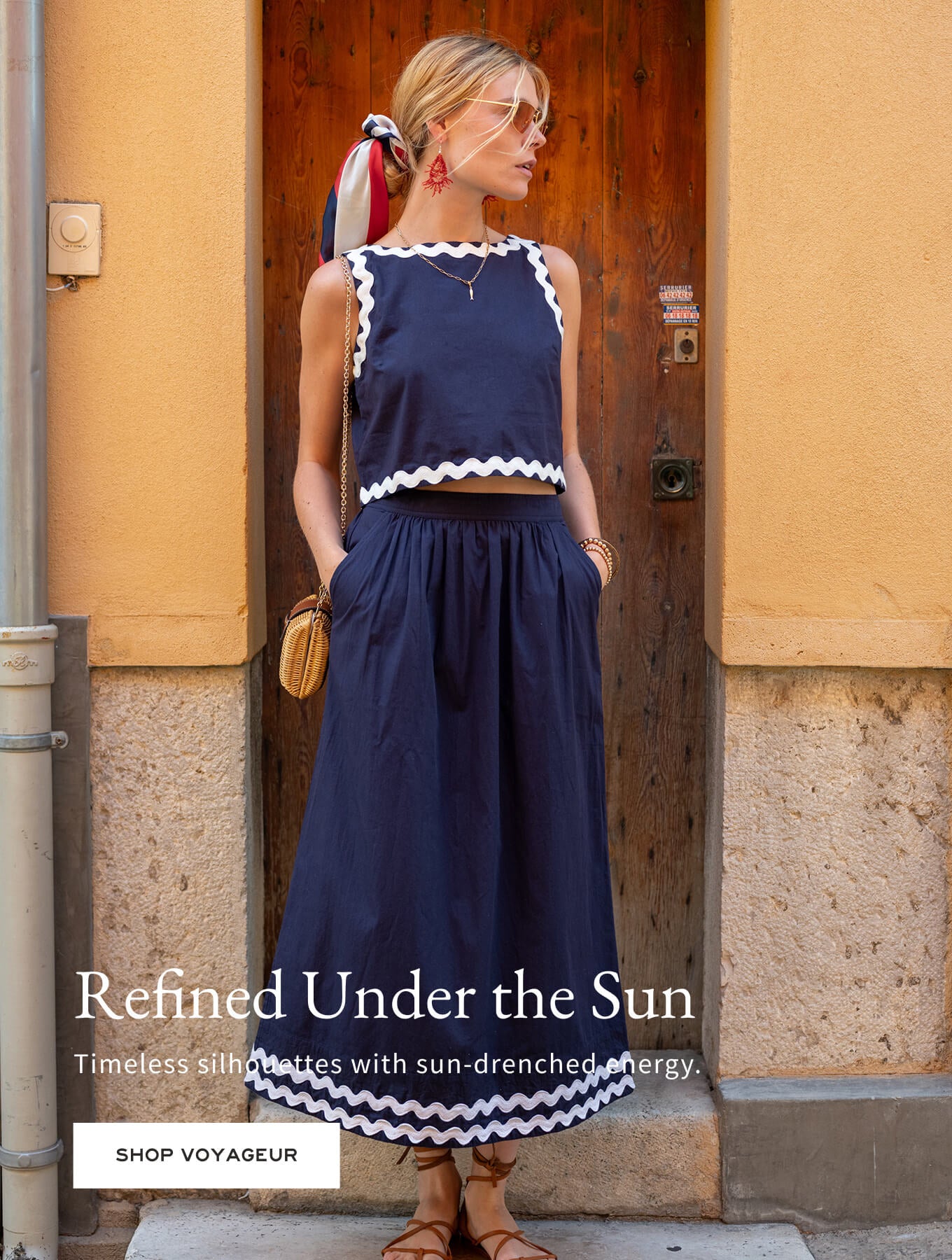 Woman in a navy dress standing in front of a wooden door with text 'Refined Under the Sun' and 'Shop Voyageur'.