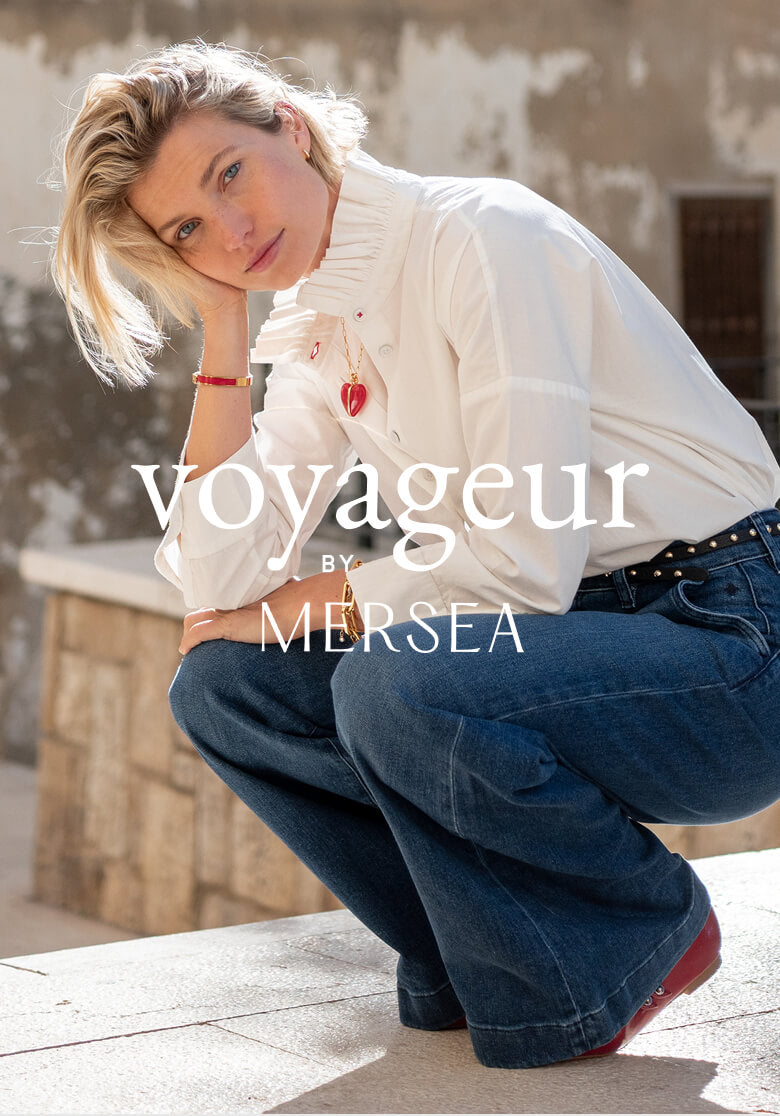 A woman in a white high-neck blouse and blue jeans posing against a rustic stone wall with the superimposed text: voyageur BY MERSEA
