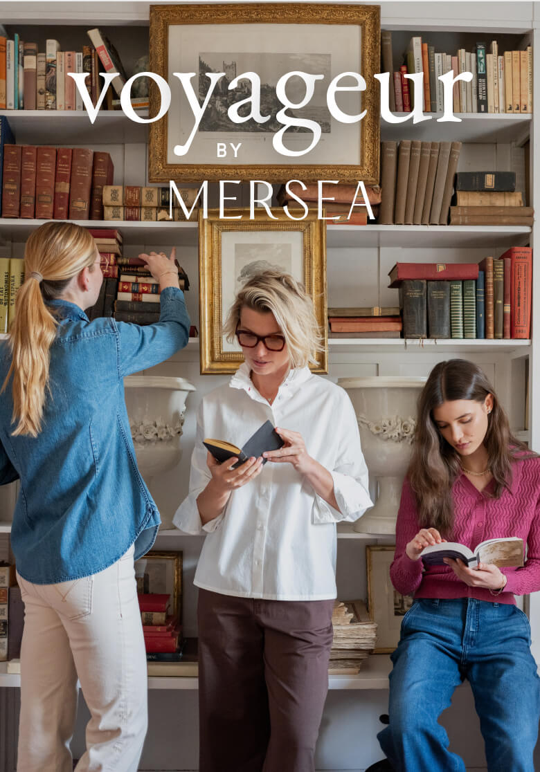 A lifestyle scene featuring three women in travel-friendly apparel in a cozy, wood-paneled library setting. One woman in a blue button-down shirt looks at a book, a middle woman in a white linen shirt and glasses reads a book, and a third woman in a pink top sits on the floor reading. Text: voyageur BY MERSEA