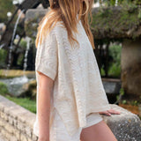 Woman wearing a light-colored lace top and shorts standing outdoors with a fountain in the background.