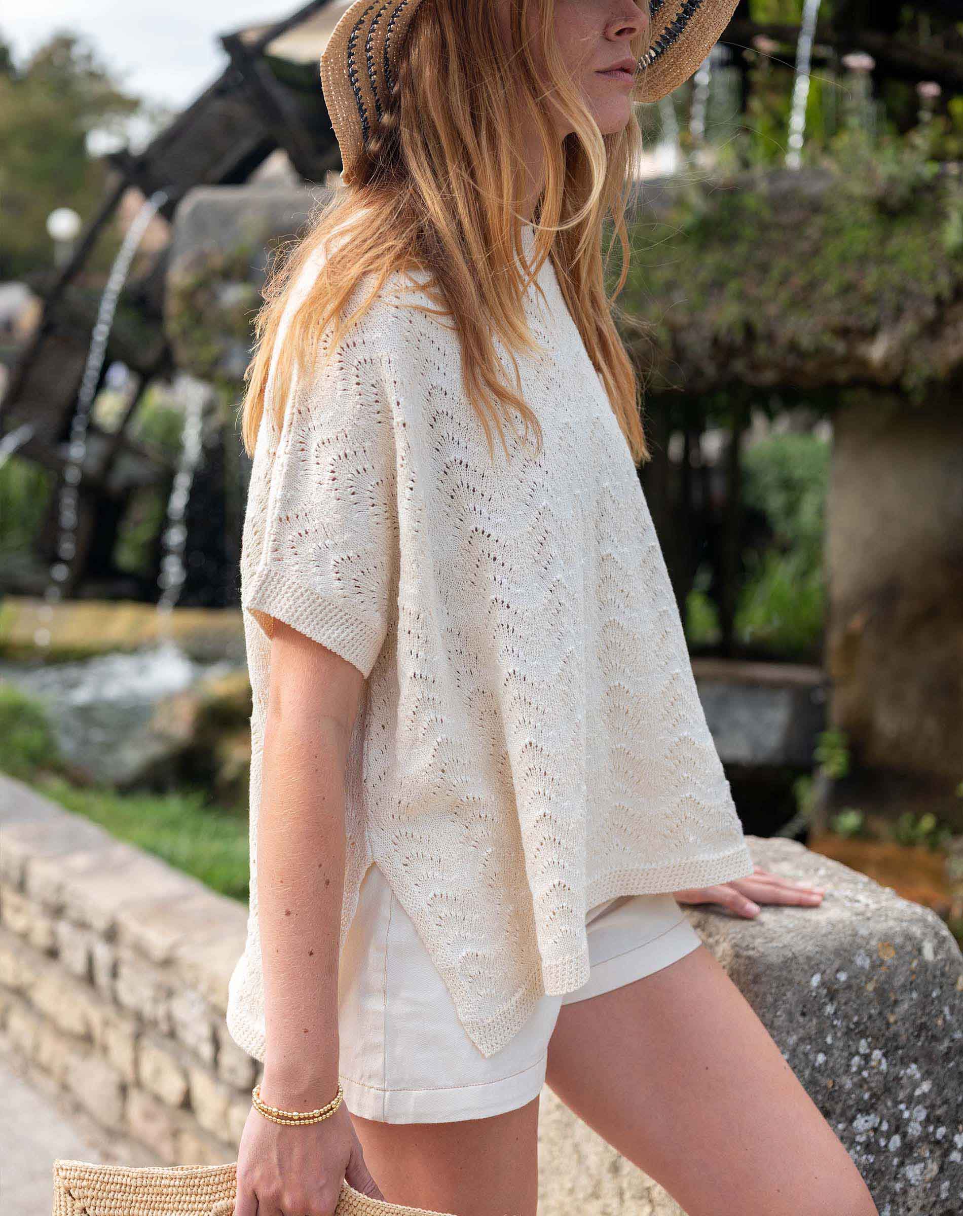Woman wearing a light-colored lace top and shorts standing outdoors with a fountain in the background.