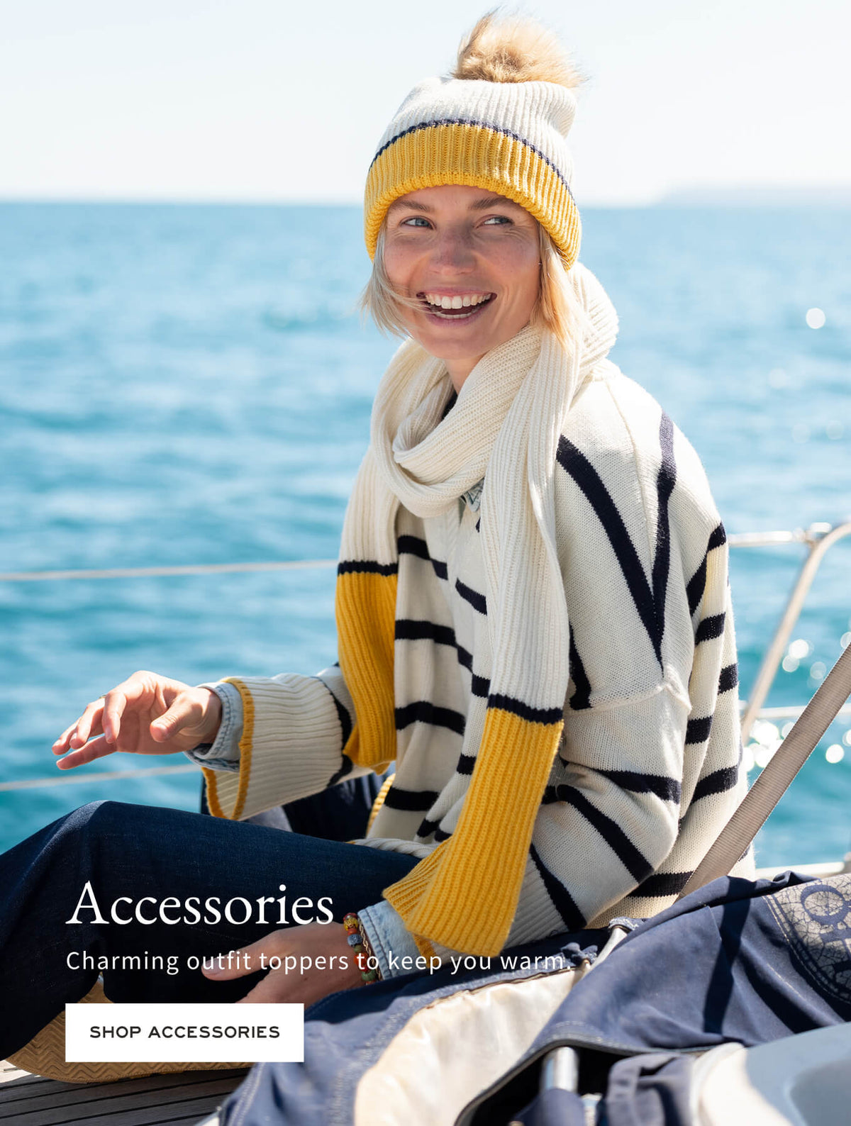 A smiling woman on a boat wearing a yellow and white striped knit sweater and pom-pom beanie. Text: 'Accessories. Charming outfit toppers to keep you warm. SHOP ACCESSORIES'
