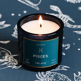 Candle in a black container labeled 'Pisces' by Mersea on a dark blue background with zodiac signs.
