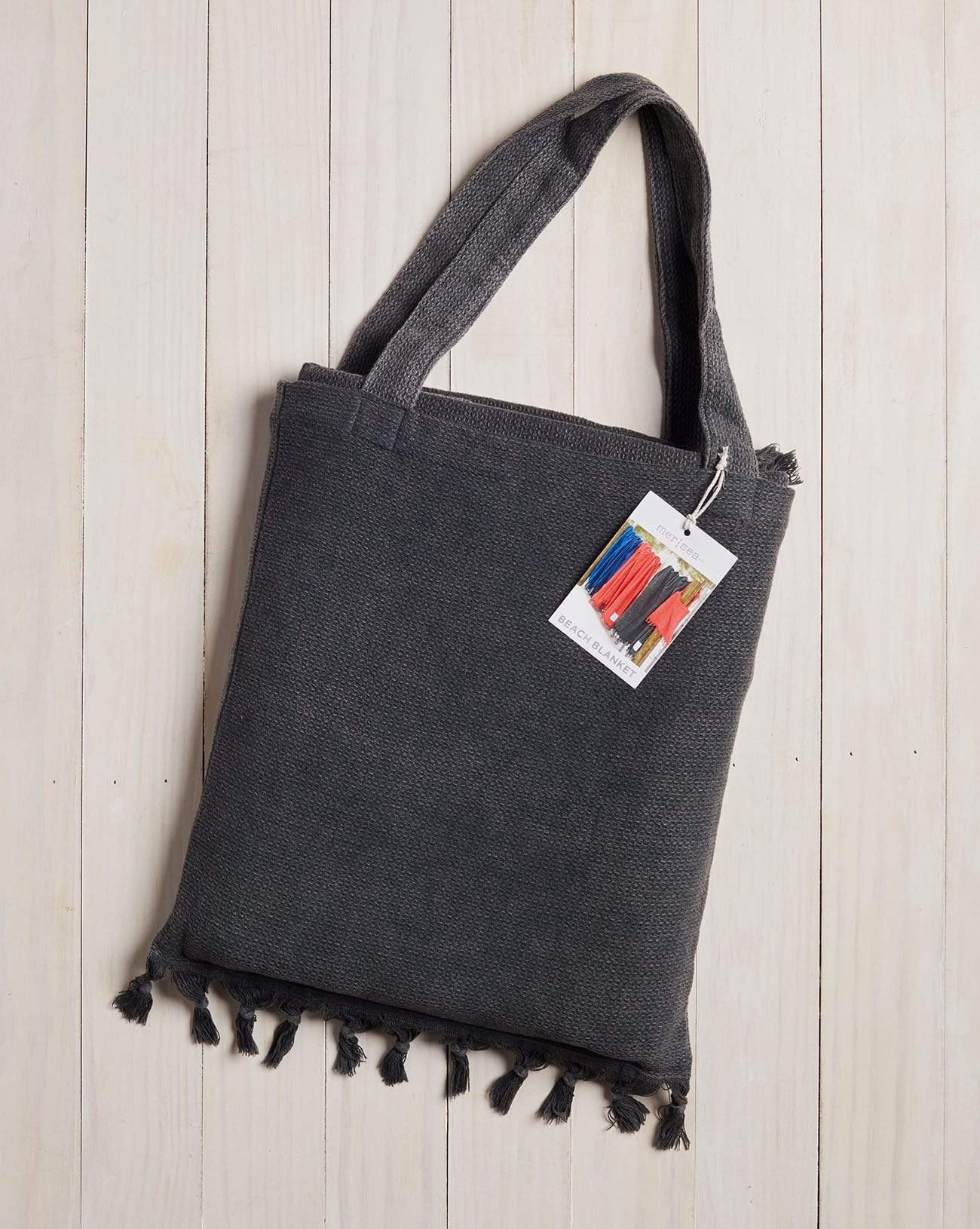 dark grey beach bag with fringe at the bottom on a white wood background