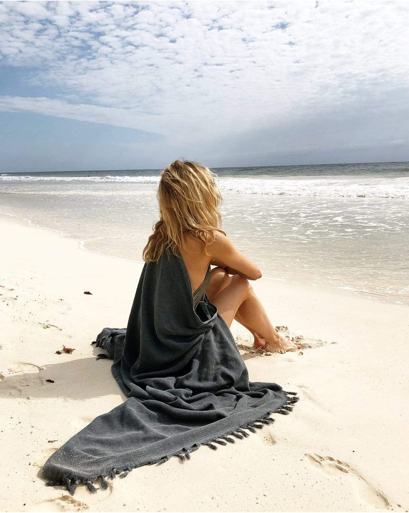 female sitting on the sand wrapped in dark grey with fringe beach blanket near the ocean