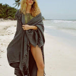 female wrapped in dark grey beach blanket with fringe walking in the san on the beach 